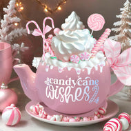 alt= A pink teapot decorated with candy canes and whipped cream, surrounded by Christmas ornaments and peppermint candies. The words 