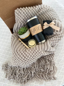 
A cozy gift set includes a textured gray blanket, a black tumbler labeled "Hello Morning" a small potted succulent, a gold tin candle, wooden matches, and a pair of dark gray socks tied with a beige ribbon.