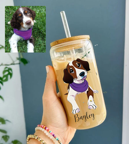 A hand holds a glass tumbler of iced coffee with a bamboo lid and glass straw. The tumbler features an illustration of a dog in a purple harness with the name 