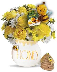 A cheerful arrangement of yellow roses, daisies, and white flowers in a honey pot vase labeled "HONEY." Accompanied by a small knitted bee, a "Bee Well Soon!" card, and a miniature beehive decoration with a bee.