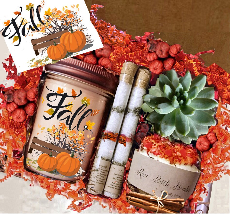
A fall-themed gift set includes a candle in a jar labeled "Fall" with pumpkin and leaf designs, two rolled bath salts, a succulent plant, a bath bomb, cinnamon sticks, and a note, all nestled in red and orange decorative filler.